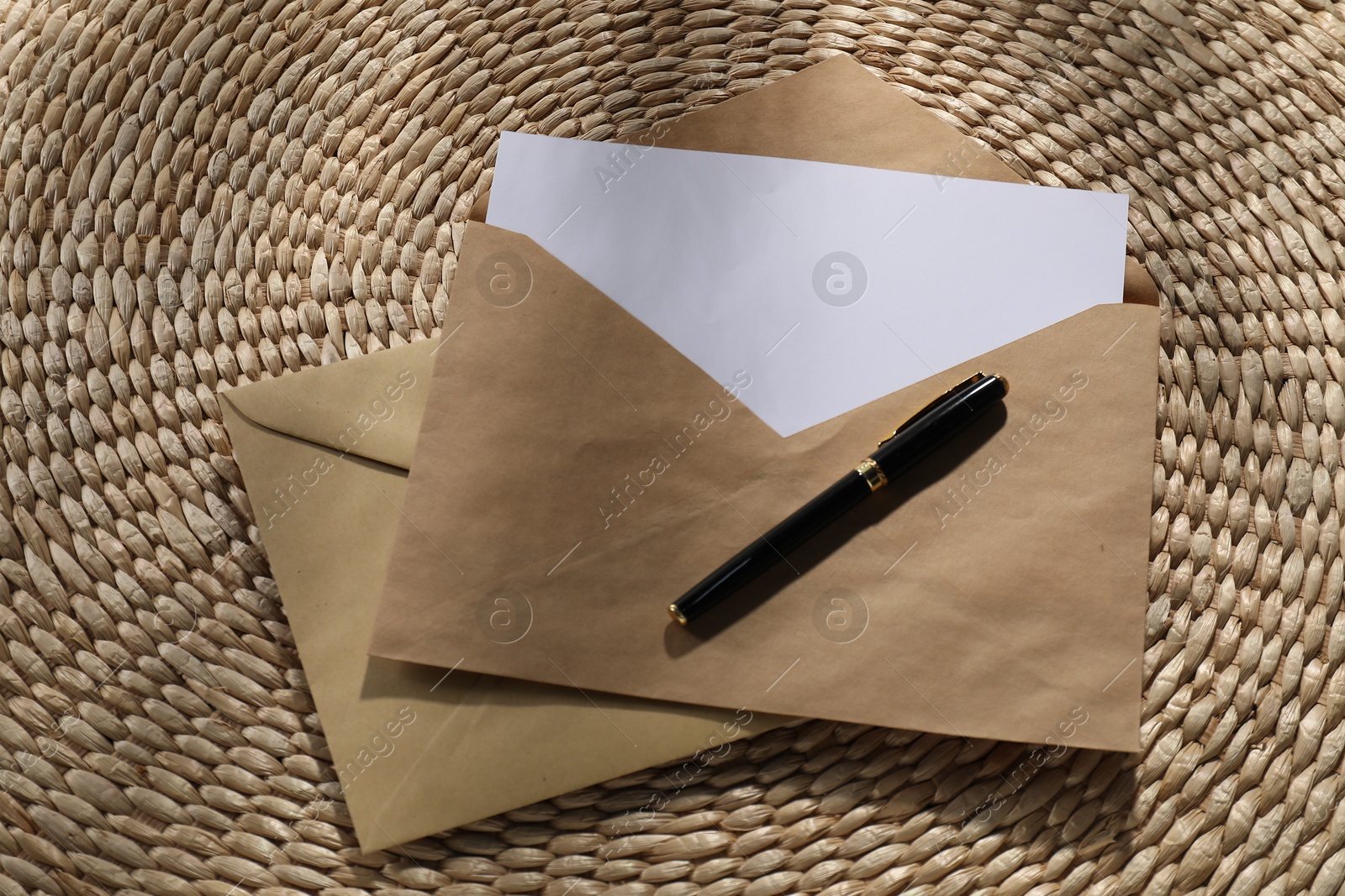 Photo of Kraft paper envelopes with letter and fountain pen on wicker mat, above view. Mockup for design