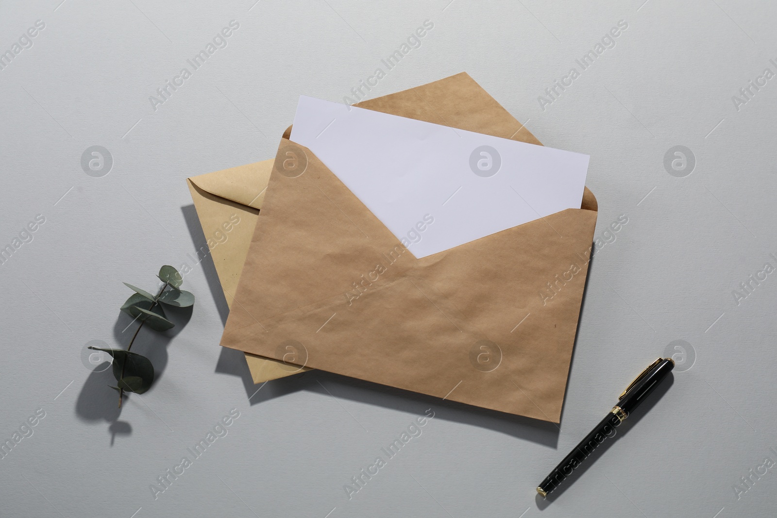 Photo of Kraft paper envelopes with letter, fountain pen and eucalyptus branch on grey background, flat lay. Mockup for design