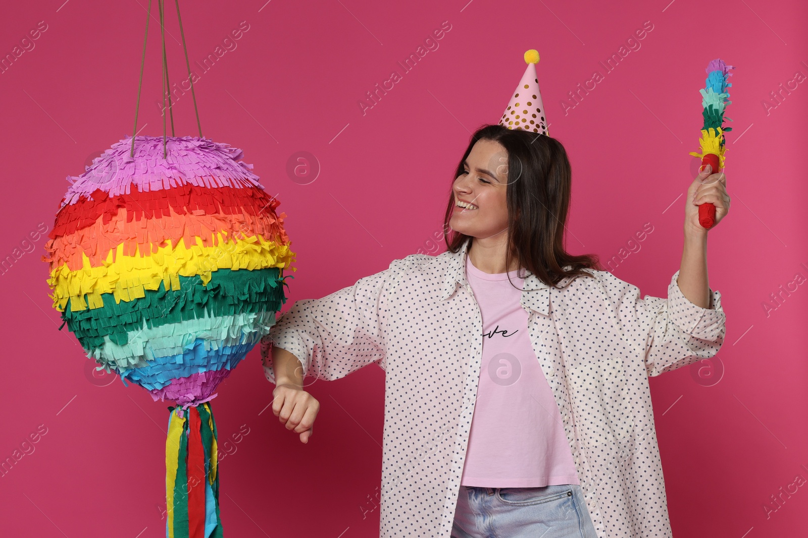 Photo of Happy woman hitting colorful pinata with stick on pink background