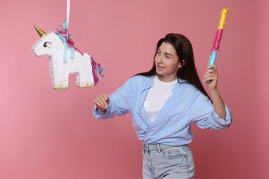 Photo of Happy woman hitting unicorn shaped pinata with stick on pink background