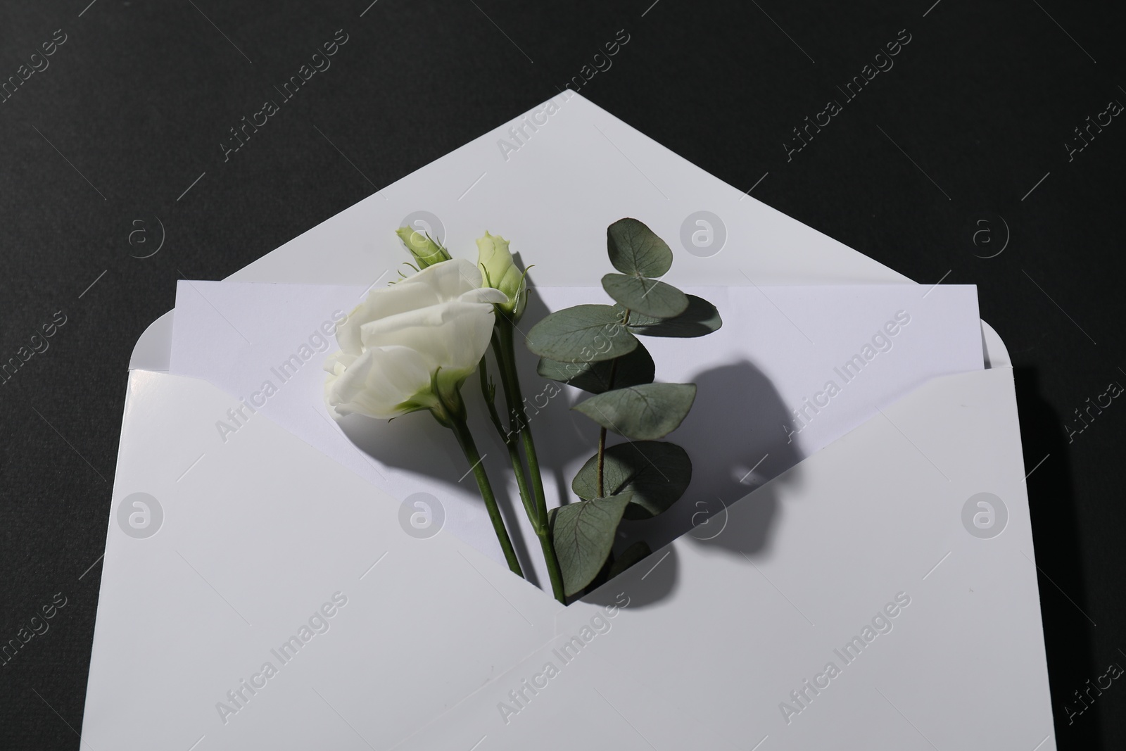 Photo of Paper envelope with letter, flowers and eucalyptus branch on black background, above view. Mockup for design