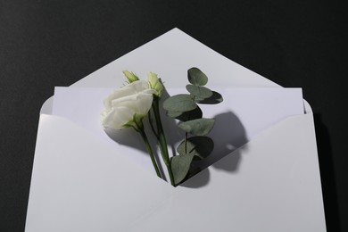 Photo of Paper envelope with letter, flowers and eucalyptus branch on black background, above view. Mockup for design