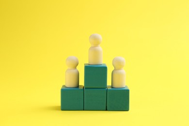 Photo of Human figures on green cubes, wooden one on top against yellow background. Competition concept