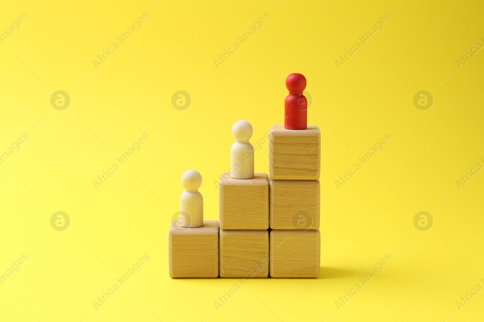 Photo of Human figures on stacked wooden cubes, red one on top against yellow background. Competition concept