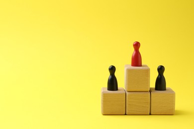 Photo of Human figures on wooden cubes, red one on top against yellow background, space for text. Competition concept