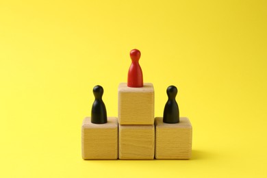 Photo of Human figures on wooden cubes, red one on top against yellow background. Competition concept