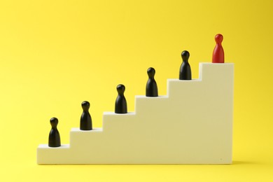 Photo of Human figures on stairs, red one on top against yellow background. Competition concept