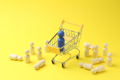 Photo of Human figures around small shopping cart with blue figure against yellow background. Competition concept