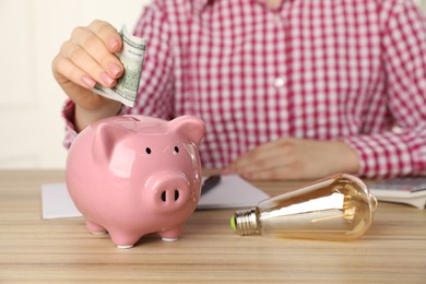 Photo of Woman putting dollar banknote into piggy bank at wooden table, closeup. Energy saving concept