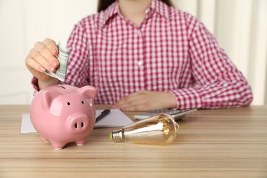 Photo of Woman putting dollar banknote into piggy bank at wooden table, closeup. Energy saving concept