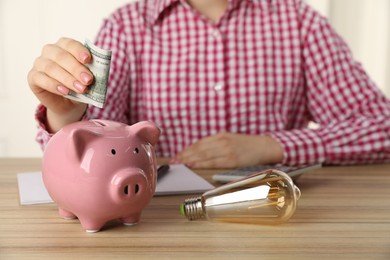 Photo of Woman putting dollar banknote into piggy bank at wooden table, closeup. Energy saving concept