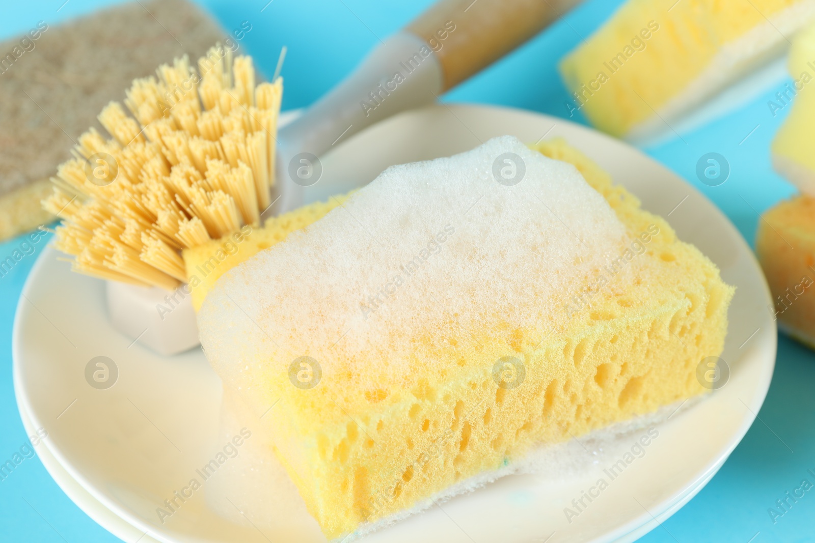 Photo of Sponge with foam, plate and brush on light blue background, closeup