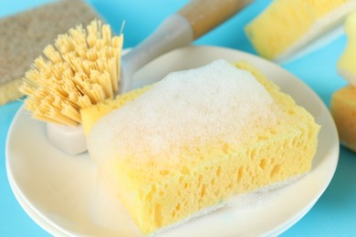 Photo of Sponge with foam, plate and brush on light blue background, closeup