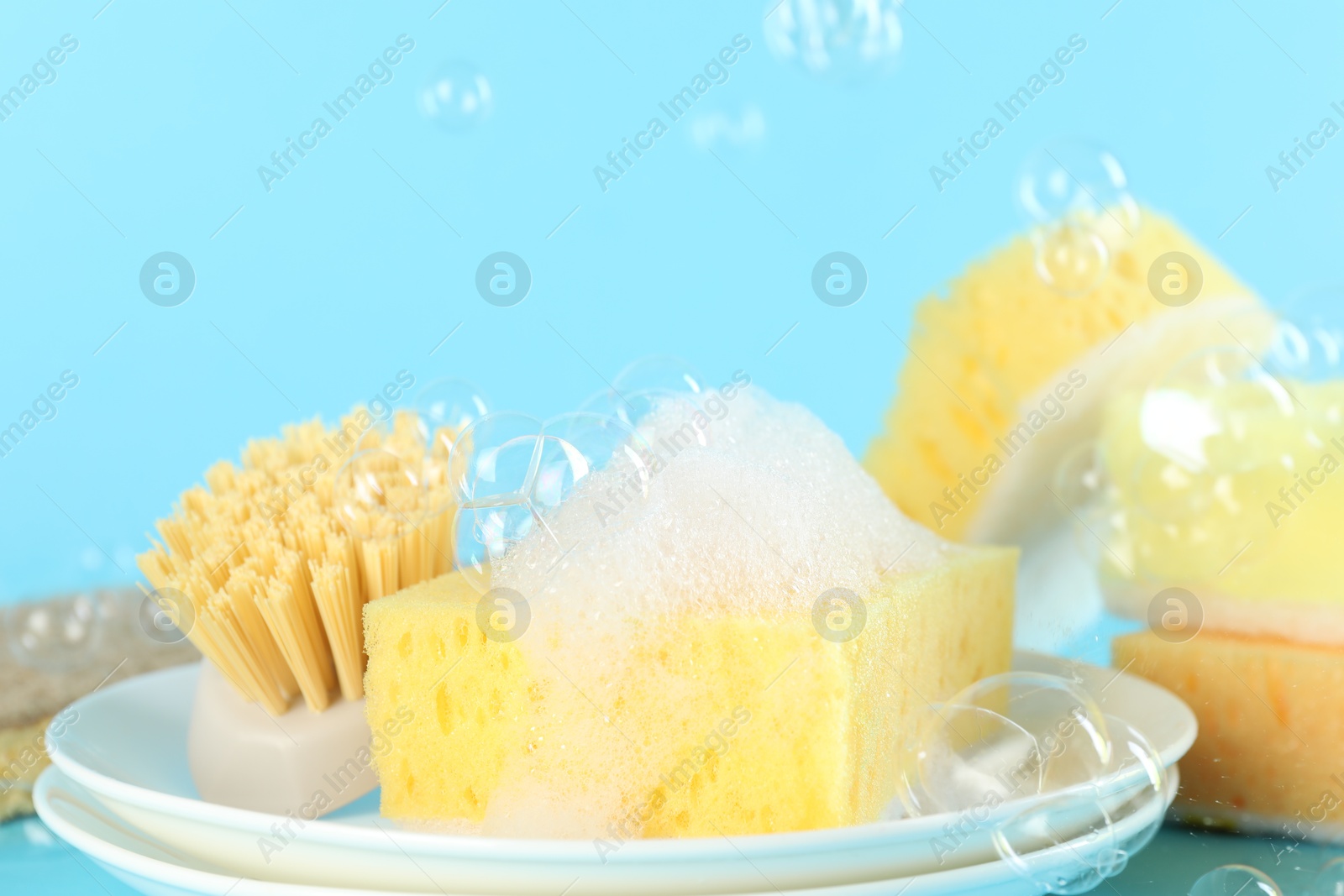 Photo of Sponges, foam, plates and brush among flying bubbles on light blue background, closeup