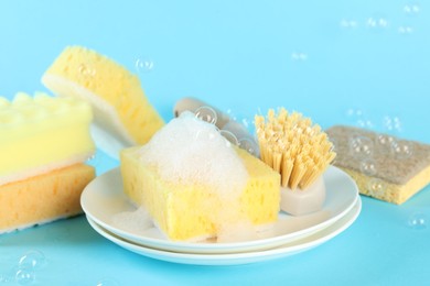 Photo of Sponges, foam, plates and brush among flying bubbles on light blue background, closeup