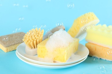 Photo of Sponges, foam, plates and brush among flying bubbles on light blue background, closeup