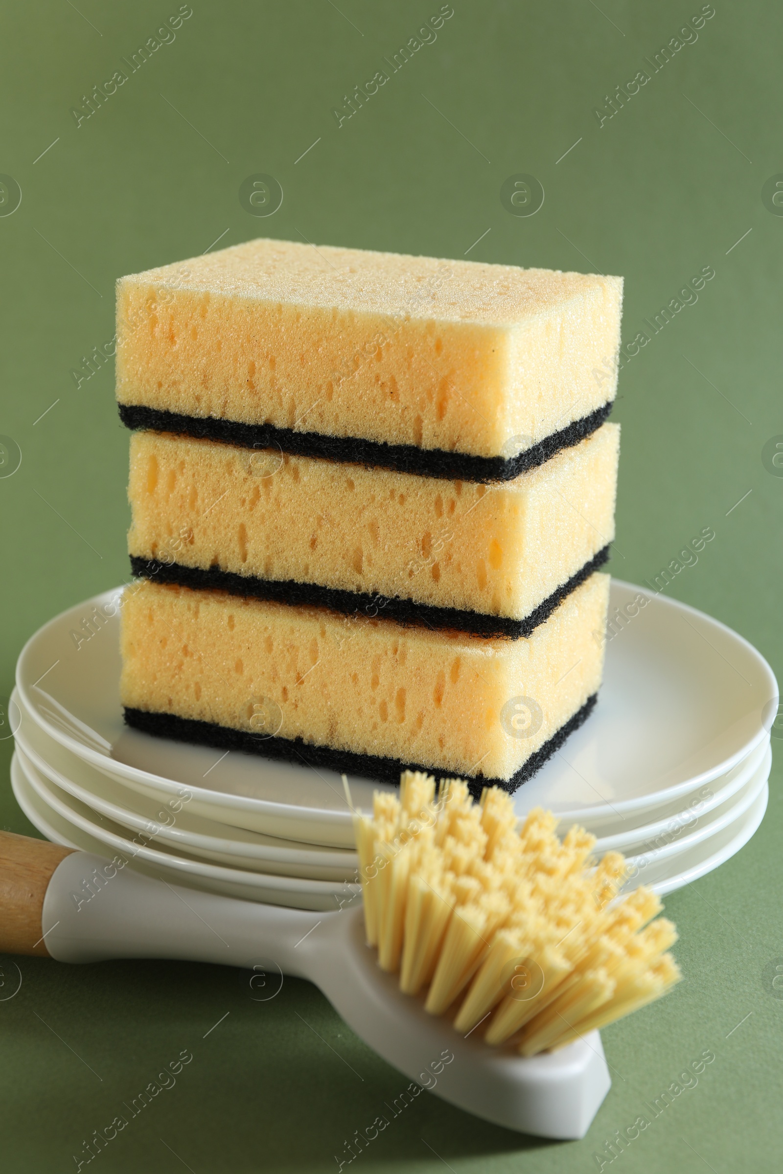 Photo of Stack of sponges, clean plates and brush on green background, closeup