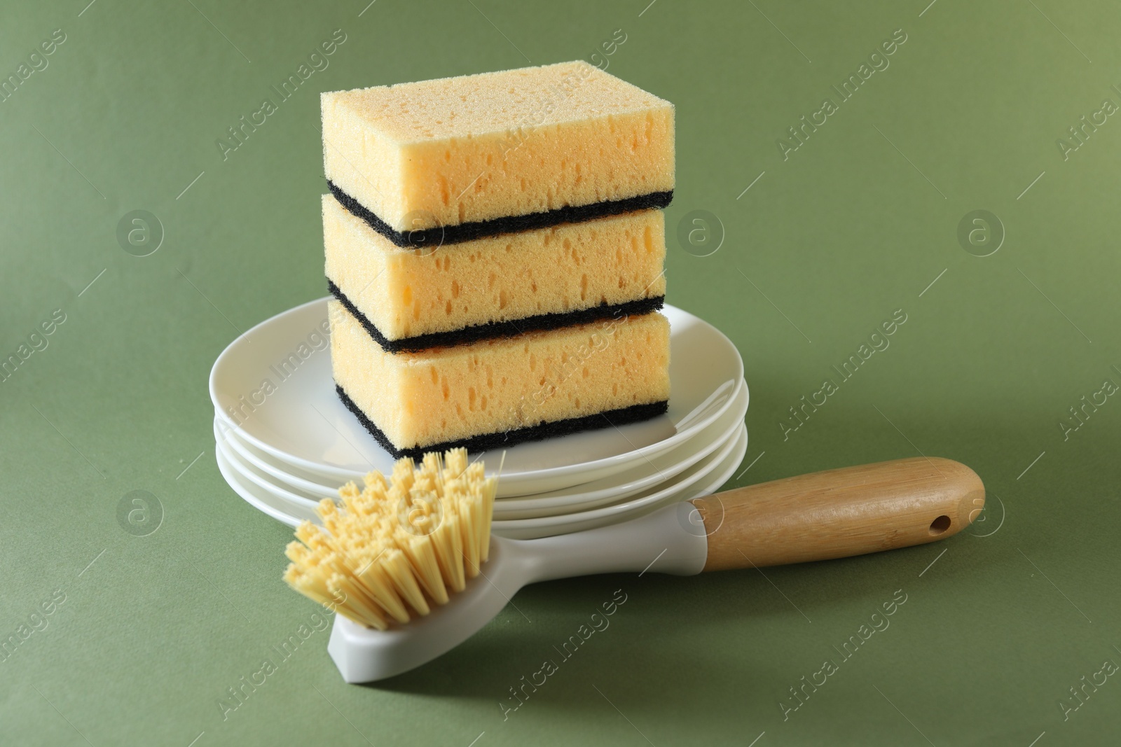 Photo of Stack of sponges, clean plates and brush on green background, closeup