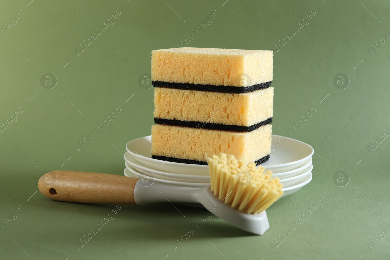 Photo of Stack of sponges, clean plates and brush on green background, closeup