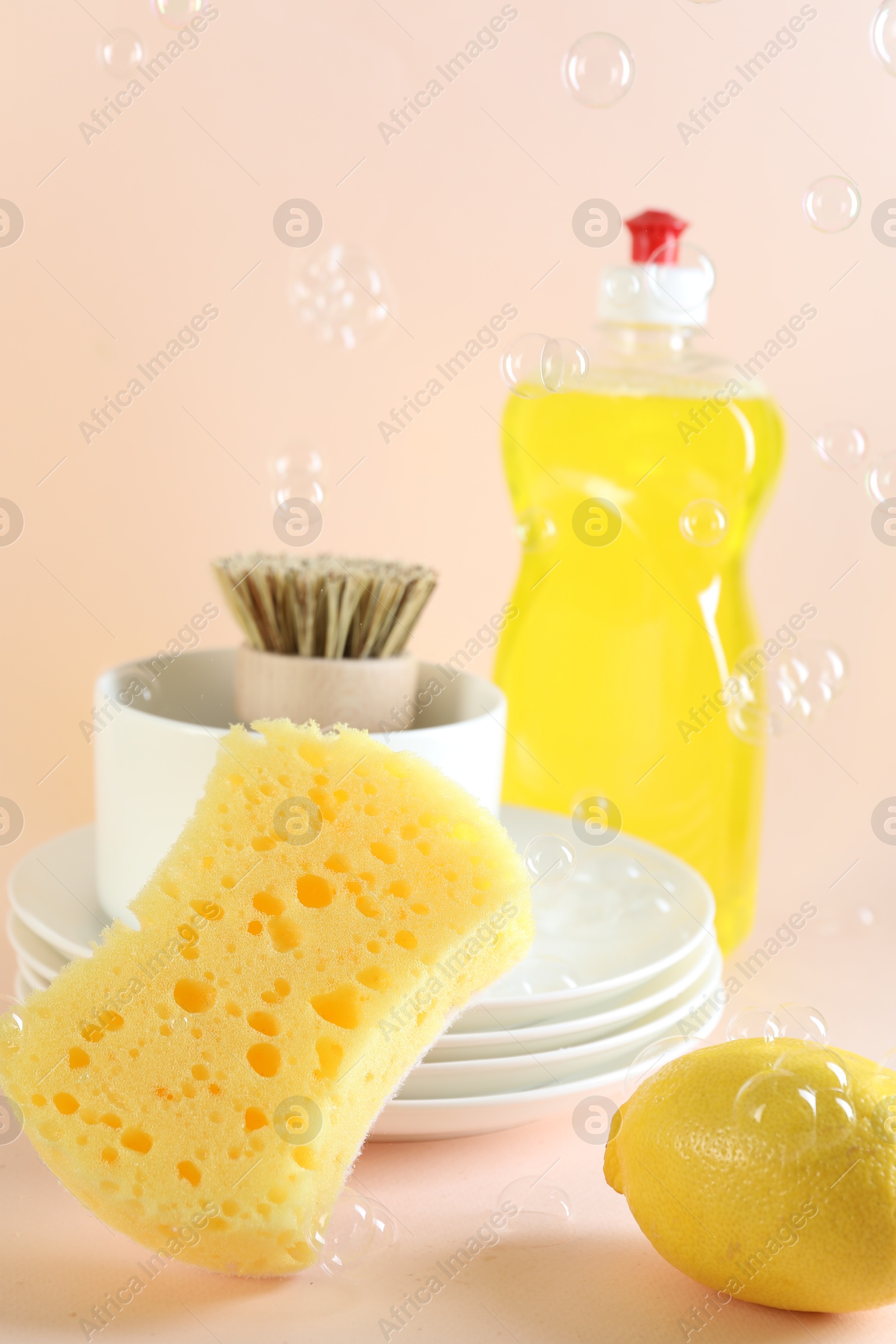 Photo of Sponge, dishware, lemon, detergent and brush among flying bubbles on beige background, closeup