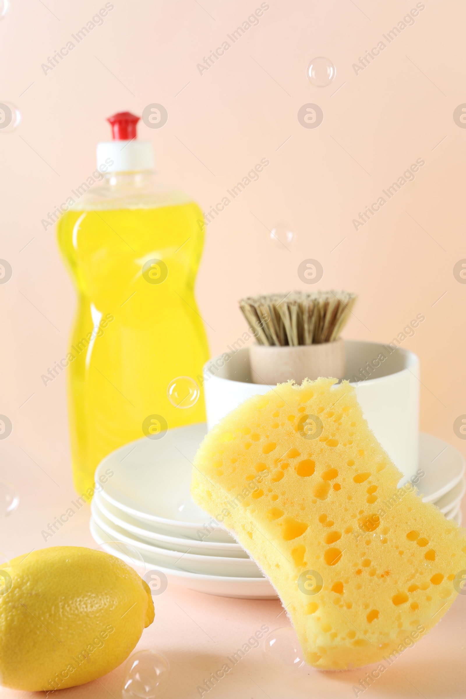 Photo of Sponge, dishware, lemon, detergent and brush among flying bubbles on beige background, closeup