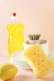 Photo of Sponge, dishware, lemon, detergent and brush among flying bubbles on beige background, closeup