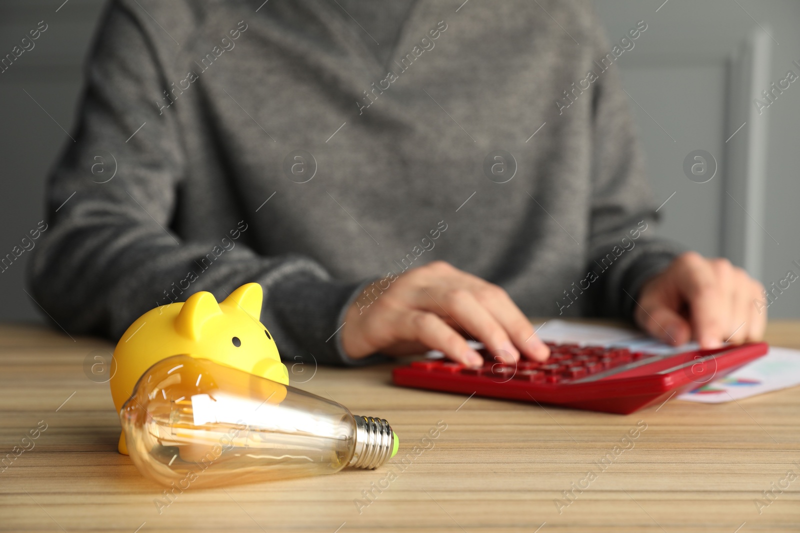 Photo of Man using calculator at wooden table, focus on piggy bank and light bulb. Energy saving concept