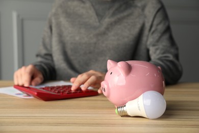 Photo of Man using calculator at wooden table, focus on piggy bank and light bulb. Energy saving concept