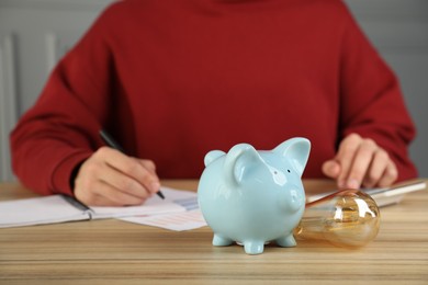 Photo of Man taking notes while using calculator at wooden table, focus on piggy bank and light bulb. Energy saving concept