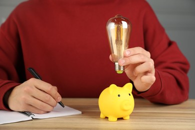 Photo of Man holding light bulb above piggy bank while taking notes at wooden table, closeup. Energy saving concept