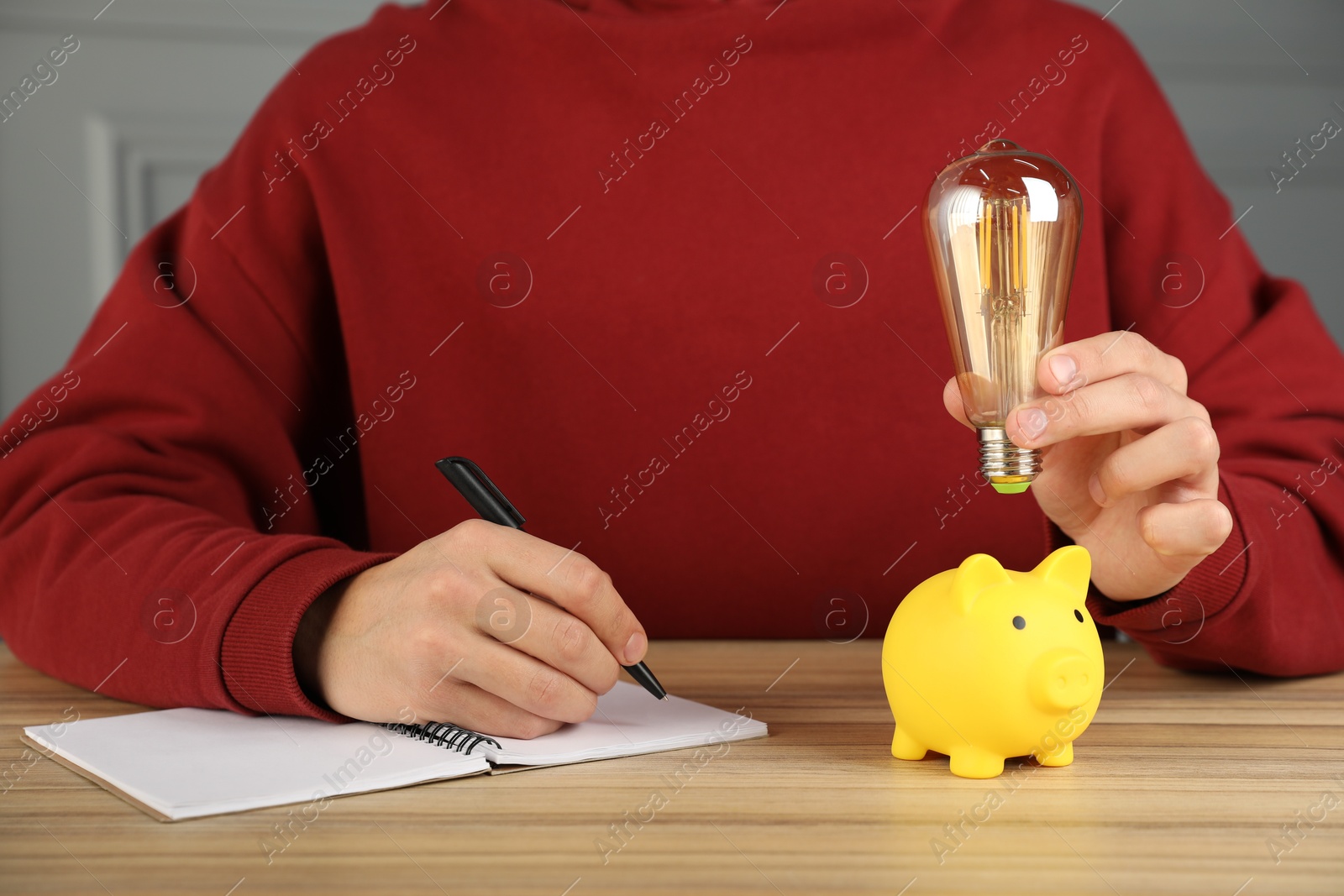 Photo of Man holding light bulb above piggy bank while taking notes at wooden table, closeup. Energy saving concept