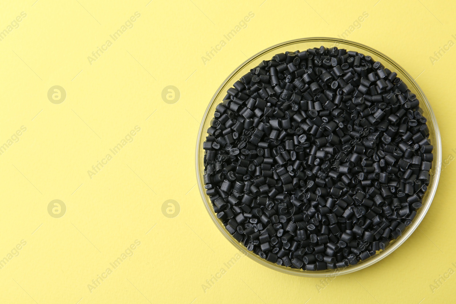 Photo of Black plastic granules in petri dish on yellow background, top view. Space for text