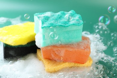 Photo of Different clean sponges with foam among flying bubbles on green background, closeup