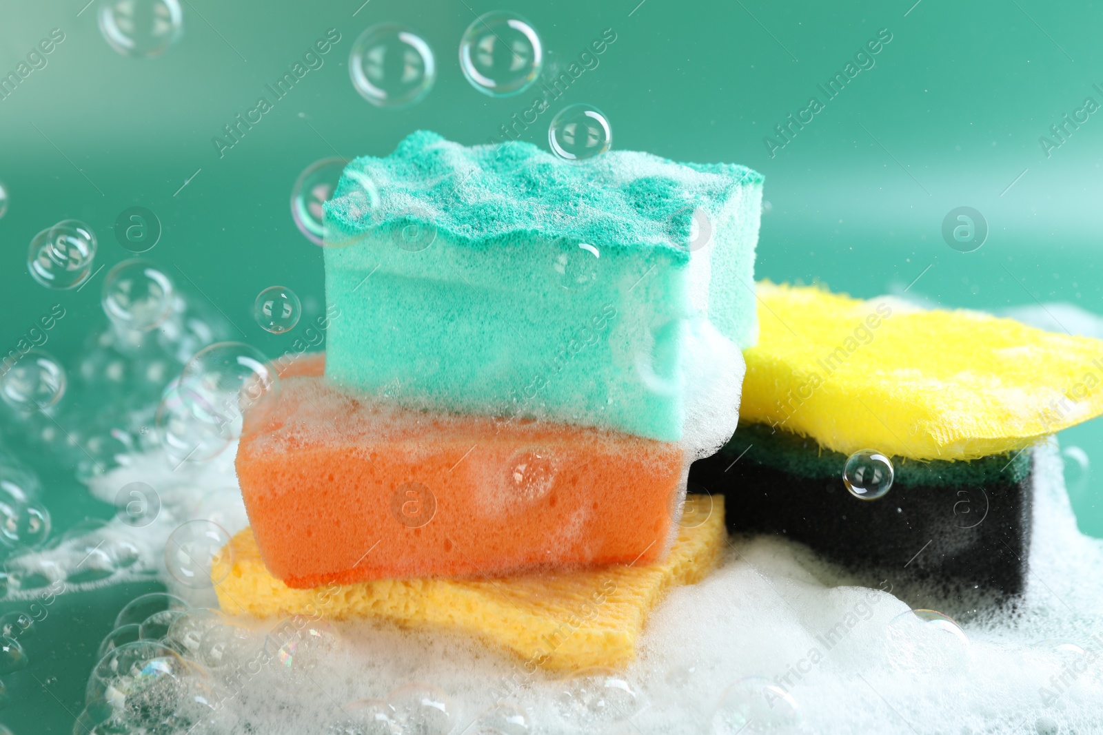 Photo of Different clean sponges with foam among flying bubbles on green background, closeup