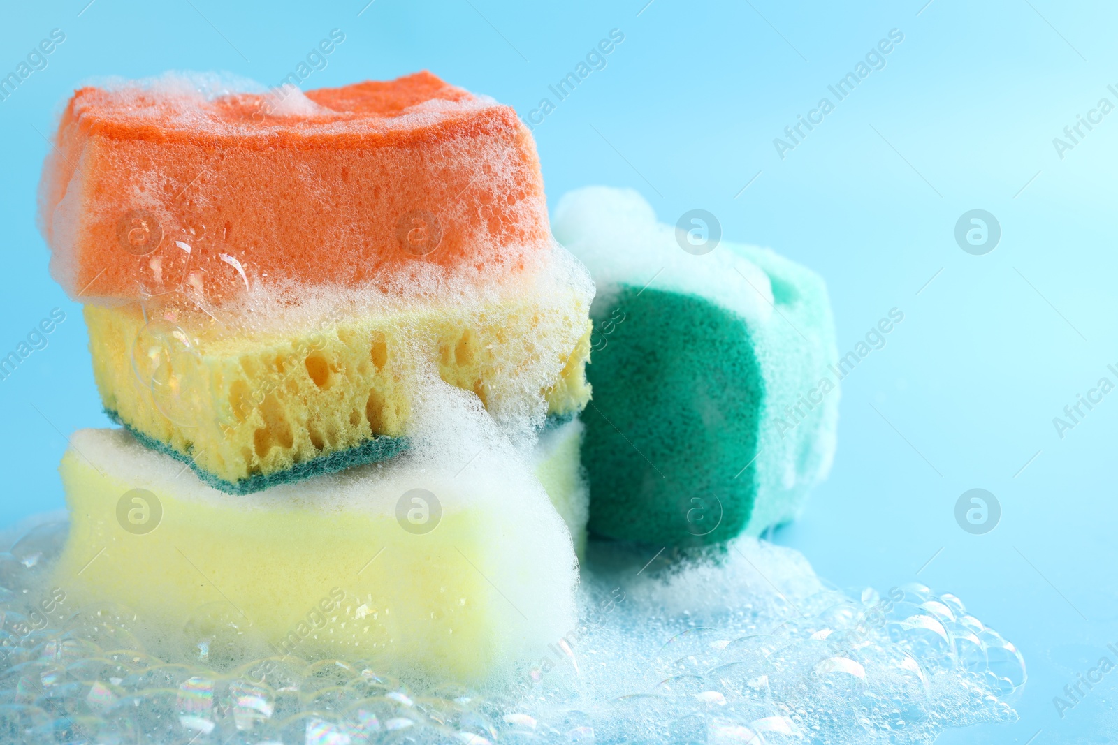 Photo of Colorful sponges with foam and bubbles on light blue background, closeup. Space for text