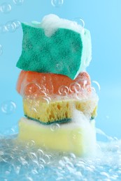 Photo of Stack of colorful sponges with foam among flying bubbles on light blue background, closeup