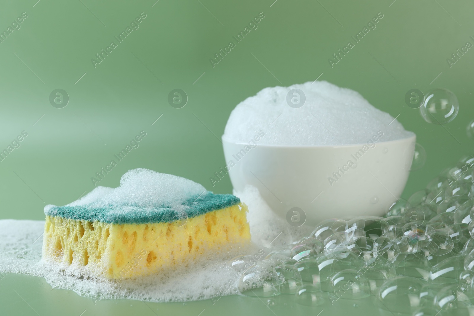 Photo of Sponge with foam, bowl and bubbles on green background, closeup