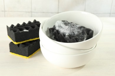 Photo of Many sponges and stack of bowls on light wooden table, closeup