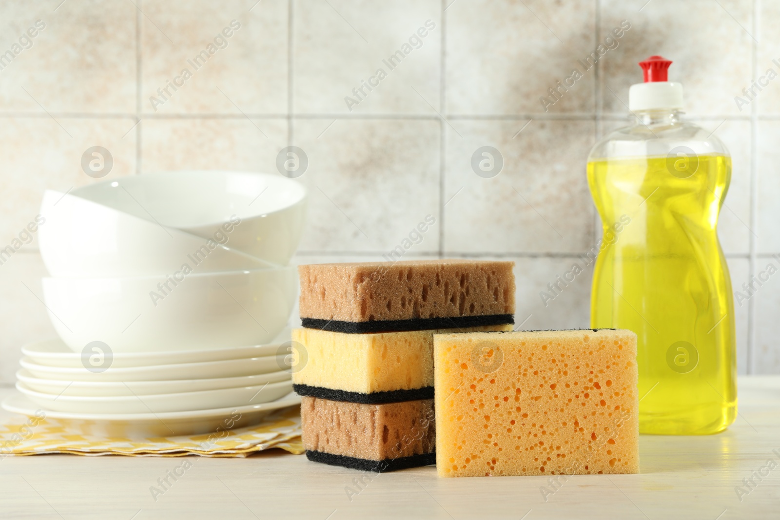 Photo of Sponges, detergent and stack of dishware on light wooden table, closeup