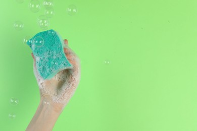 Photo of Woman holding sponge with foam among flying bubbles on green background, closeup. Space for text