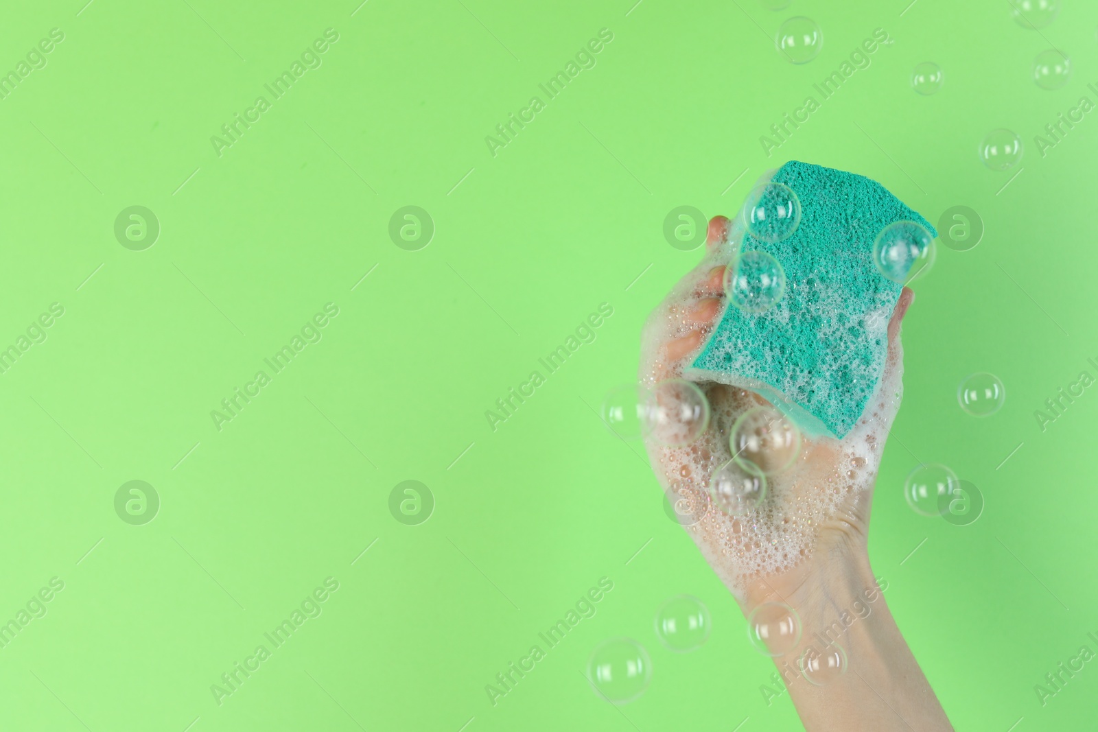 Photo of Woman holding sponge with foam among flying bubbles on green background, closeup. Space for text