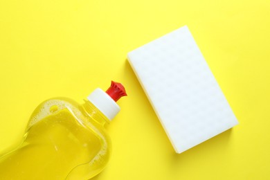 Photo of Sponge and bottle with detergent on yellow background, top view