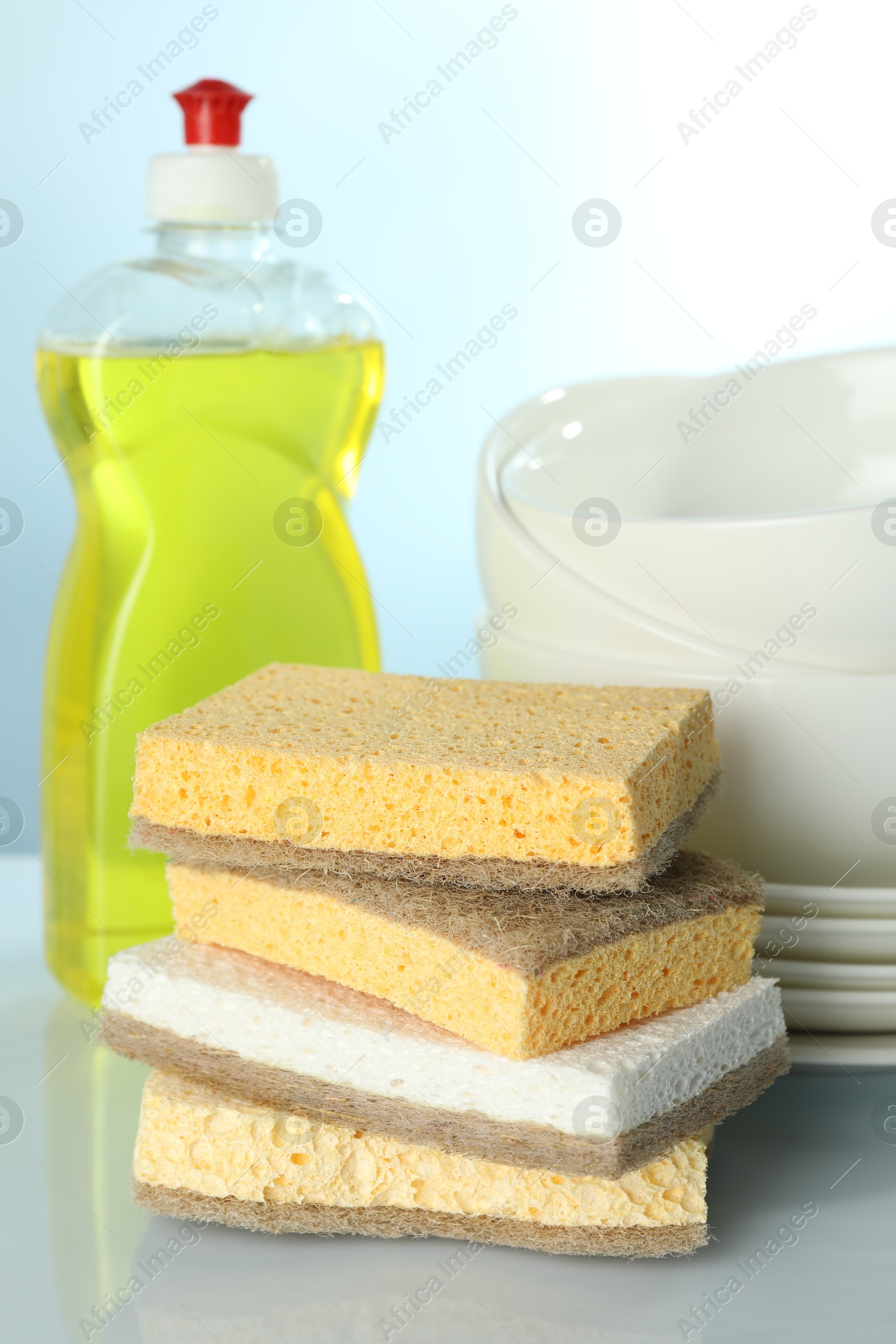 Photo of Stack of sponges, bottle with detergent and dishware on light table, closeup