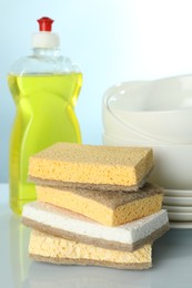 Photo of Stack of sponges, bottle with detergent and dishware on light table, closeup