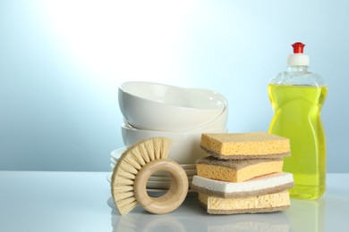 Photo of Stack of sponges, brush, bottle with detergent and dishware on light table, closeup. Space for text