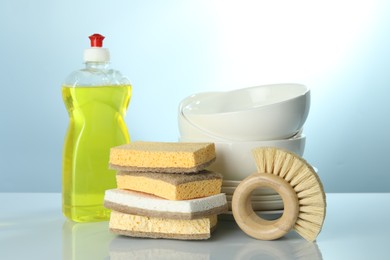 Photo of Stack of sponges, brush, bottle with detergent and dishware on light table, closeup