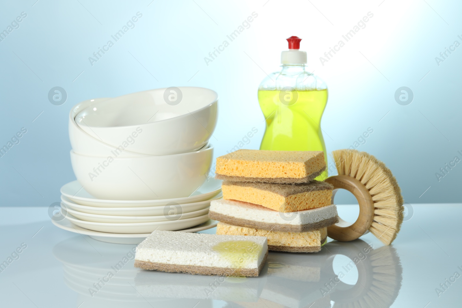 Photo of Stack of sponges, brush, bottle with detergent and dishware on light table, closeup