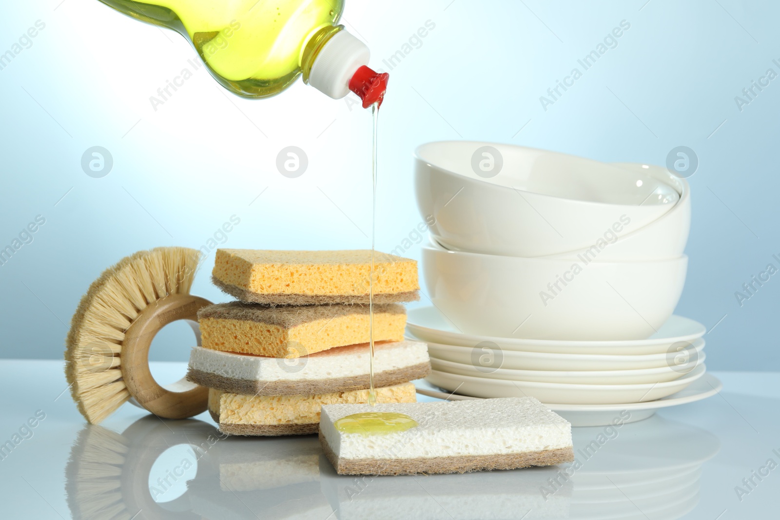 Photo of Pouring detergent from bottle onto sponge at light table with brush and dishware, closeup