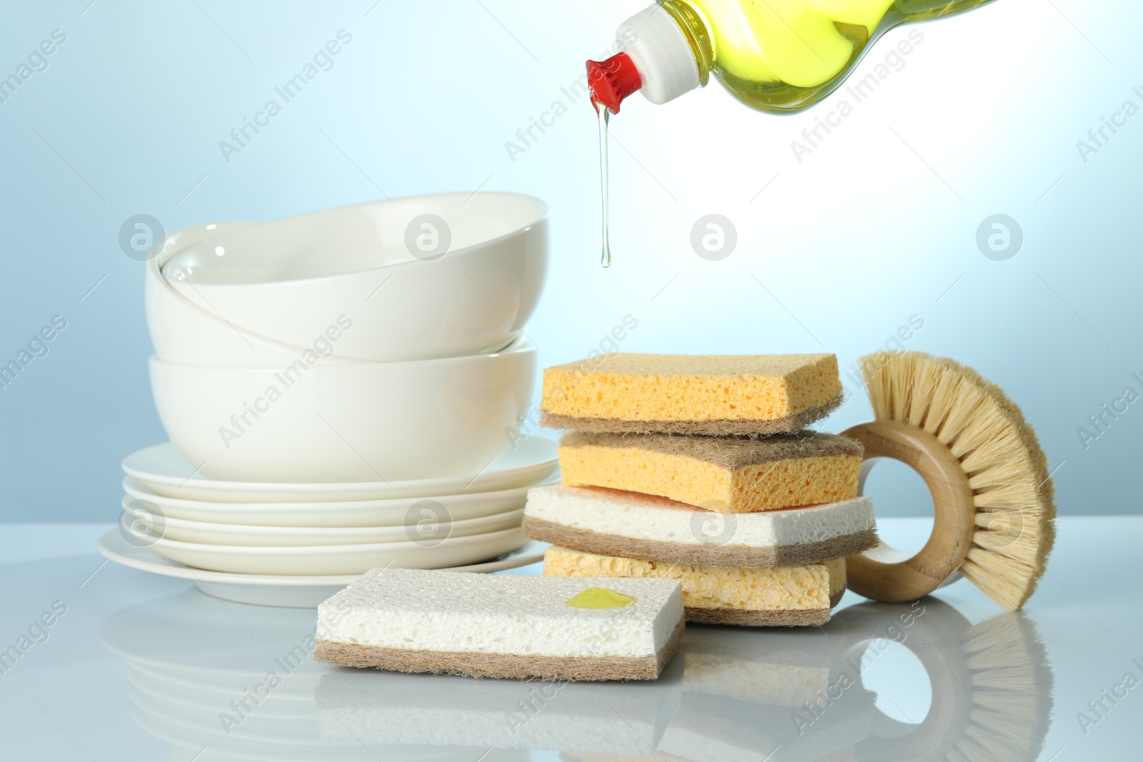 Photo of Pouring detergent from bottle onto sponge at light table with brush and dishware, closeup