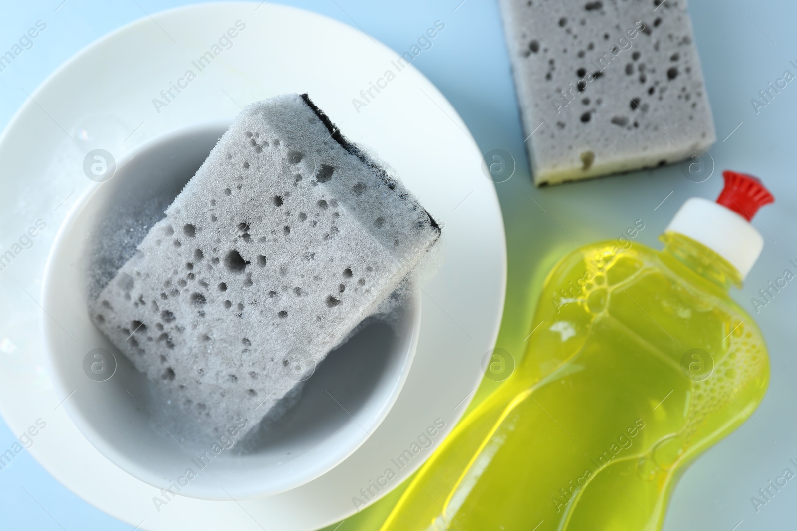 Photo of Sponges, foam, dishware and bottle of detergent on light blue background, flat lay